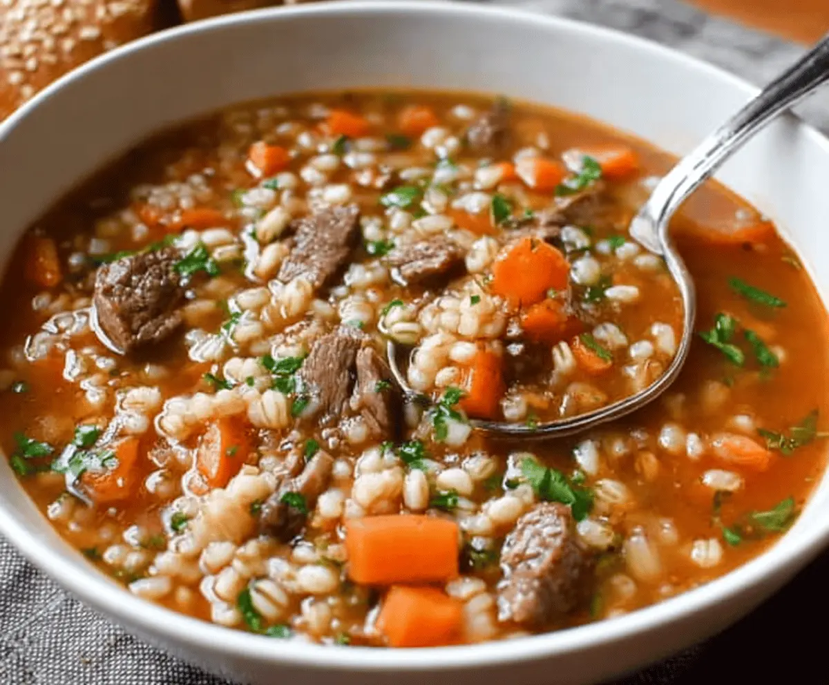 Hearty beef and barley soup in a bowl garnished with fresh herbs, served with crusty bread on a rustic wooden table.